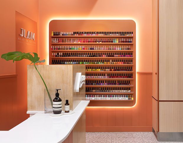 A brightly illuminated nail salon reception and retail display area. A large wall-mounted shelving unit densely stocked with colourful nail polish bottles is framed by warm LED lighting against a terracotta-orange wall. In the foreground is a white reception counter with a timber-faced built-in section, sink or basin fixtures, soap dispensers, and a leafy plant. Pale timber cabinetry and a vertical slatted or grooved panel appear at the right, while terrazzo flooring is visible below. The space has a polished, contemporary commercial interior with warm, saturated colours and high visual contrast.