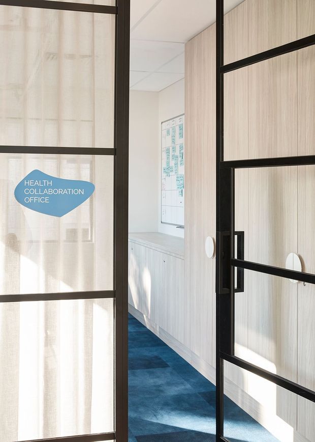 View through black-framed glazed doors into a bright health collaboration office. The space features light wood-grain wall panelling and fixed cabinetry, a blue carpeted floor, a white ceiling, and a wall-mounted planning board. Sheer curtains and strong daylight create an airy commercial workplace setting.