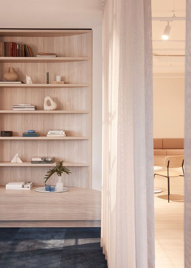 Warm, softly lit interior view featuring a full-height light timber built-in shelving and cabinetry wall with books and restrained decorative objects. Sheer curtains partially screen an adjacent sitting or dining area with a tan upholstered bench, light timber flooring, and a simple timber chair. The design is calm and minimalist with natural materials and muted neutral tones.