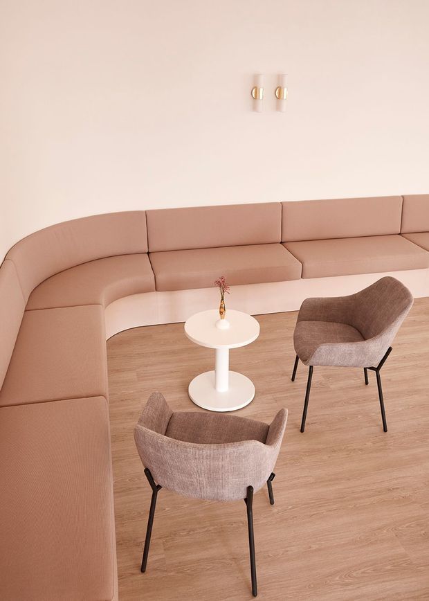 A softly styled lounge or reception area with a large curved dusty-rose upholstered banquette wrapping around the room, two textured brown armchairs, a small round white pedestal table, pale timber-look flooring, and minimal wall-mounted brass-and-white lighting. The palette is warm, muted, and refined.