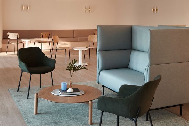 A softly lit contemporary lounge and dining area with a muted beige and brown palette. The space features a large blue-grey upholstered high-back booth or acoustic seating element, dark green lounge chairs, a round timber coffee table, pale blue-grey rug, built-in banquette seating, small round dining tables, and minimalist pendant wall lights.
