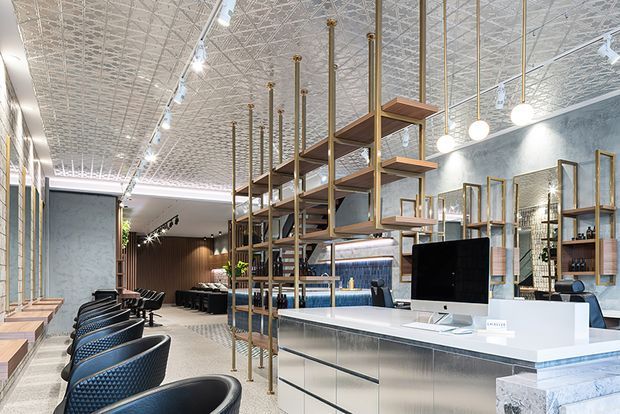 A bright, open commercial salon or showroom interior with a long white reception counter in the foreground, black salon chairs along the left side, and extensive open timber-and-brass shelving suspended from the ceiling. The space features patterned pressed-metal ceiling panels, track lighting and globe pendant lights, dark timber wall panelling, concrete-look finishes, and a polished terrazzo-style floor. The design has a refined contemporary industrial character.