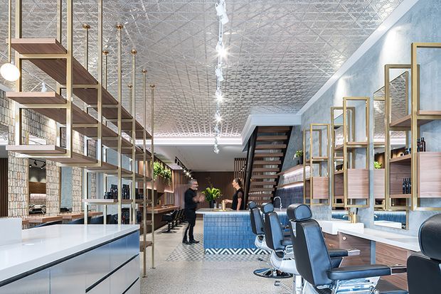 A bright, long commercial salon interior with barber or hairdressing stations along the right side, open shelving and product displays, a tiled service counter in the centre, and a patterned pressed-tin ceiling. The space combines pale blue walls, timber joinery, brass-toned frames and fittings, dark upholstered salon chairs, and patterned tile detailing. A stairway and additional seating or service area extend toward the rear.