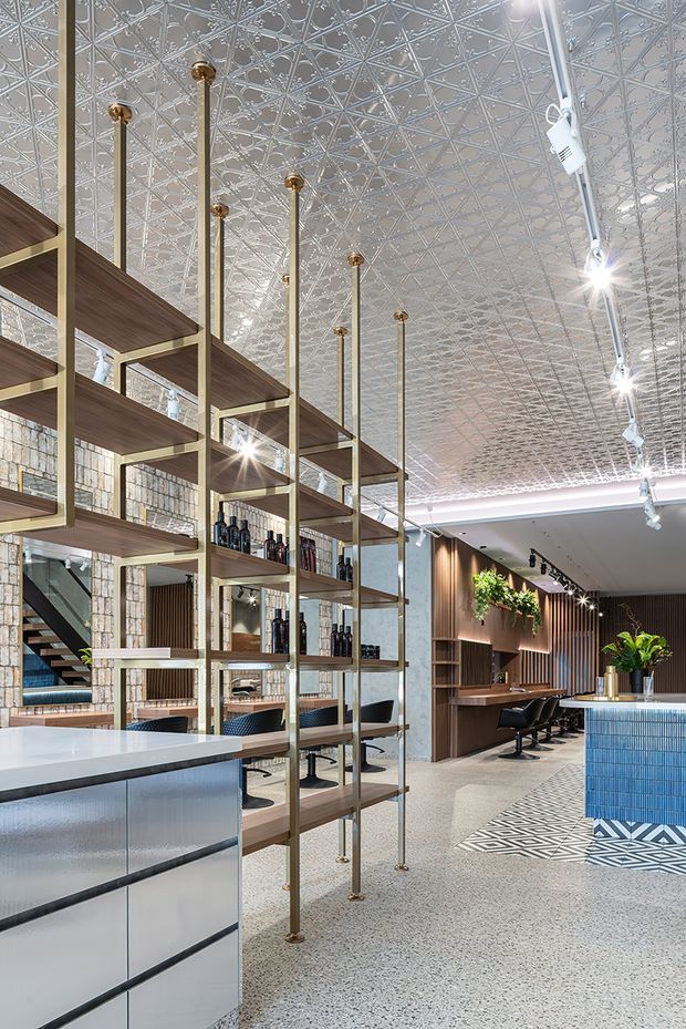 A spacious, refined commercial interior resembling a boutique salon or hospitality venue. The room features an ornate pressed-metal patterned ceiling, polished brass-toned shelving supports with open timber shelves, exposed textured masonry walls, terrazzo-style flooring, timber wall panelling, integrated workstations with salon chairs, and a blue tiled counter or feature island. Track lighting and abundant indoor planting add contemporary character.
