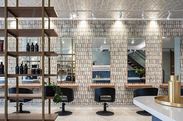 A refined commercial hospitality interior, likely a bar or lounge, with a terrazzo floor, textured light brick feature wall, timber open shelving displaying bottles, mirrored openings, and a curved white counter with brass-toned fixtures. Three dark upholstered bar or salon-style chairs sit along the feature wall. Decorative pressed-pattern ceiling panels and track lighting add visual detail, while the reflected space reveals additional seating and a stair or circulation area.