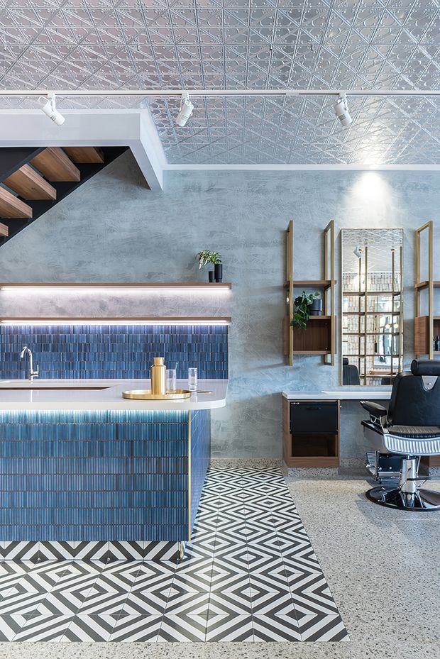 A stylish contemporary hair salon or barber shop interior with a blue tiled service counter and matching backsplash, patterned geometric floor tiles, pale textured walls, terrazzo flooring, and a decorative pressed-metal ceiling. A black barber chair sits beside timber shelving and a large mirror, while exposed timber stair treads and track lighting add architectural detail.