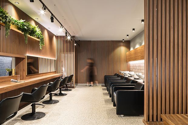 A warmly lit contemporary hair salon with a long timber styling counter and mirrors on the left, rows of black salon chairs on both sides, vertical timber slat wall cladding, integrated shelving, hanging greenery, track lighting, and a polished terrazzo-like floor. The space has an elongated open-plan layout and a refined, modern commercial fit-out.