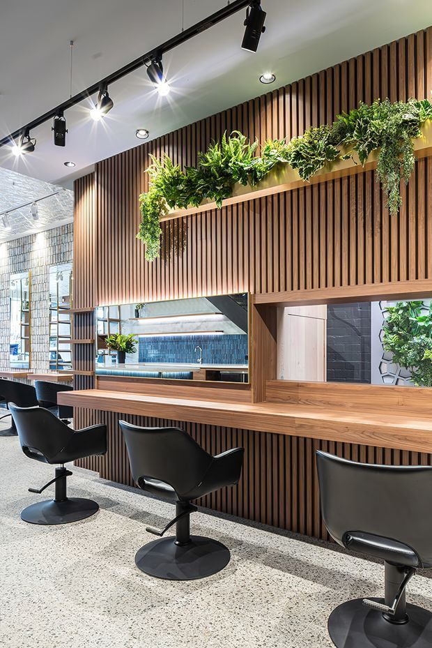 A bright contemporary commercial salon or beauty studio interior with a long timber service counter and multiple black styling chairs. The main wall is clad in vertical timber battens, with integrated mirrors and shelving displaying lush hanging greenery. Track lighting and recessed downlights illuminate the space, while a tiled service area is visible through the openings behind the counter. The floor has a light terrazzo-like finish.