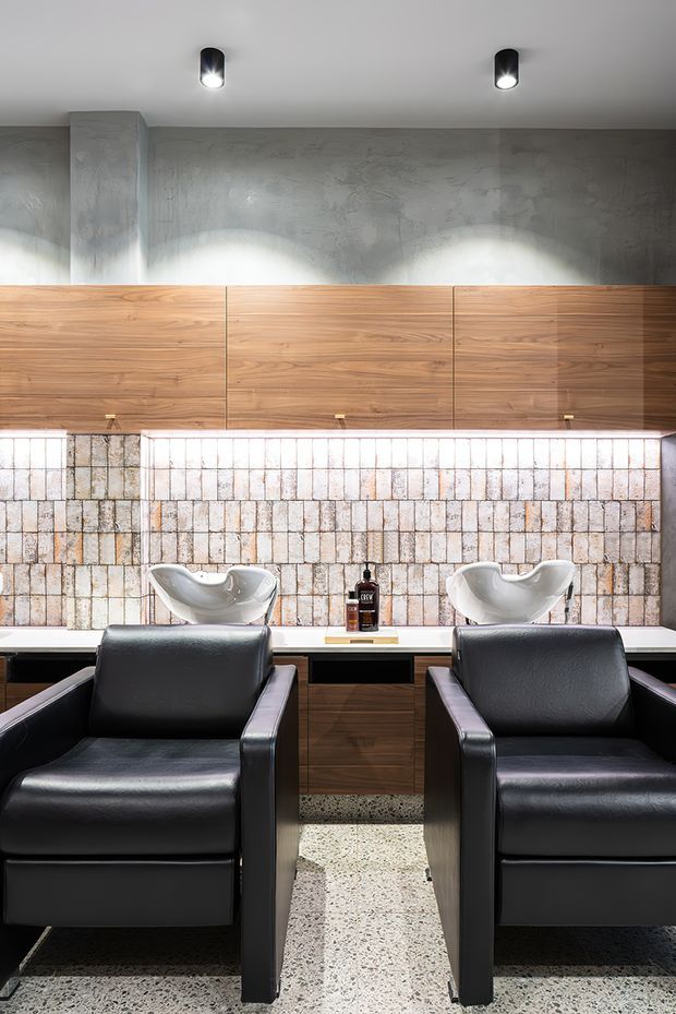 Contemporary salon interior with two black leather styling chairs facing a long counter and twin white basin fixtures. The space features a textured grey wall, extensive warm timber cabinetry, a backlit pale brick or stone-tile backsplash, and a terrazzo floor, with focused cylindrical ceiling spotlights.