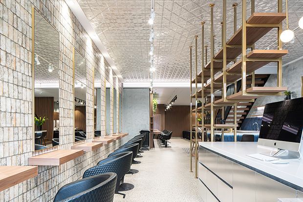 A bright, elongated commercial salon interior with multiple styling stations along a tiled feature wall and large mirrors. Black salon chairs sit at timber-topped work counters, while a white reception or service counter occupies the foreground beneath open timber shelving supported by slender brass-toned posts. The space has a decorative pressed-tin ceiling, polished light terrazzo-like flooring, track lighting, and dark timber wall accents.