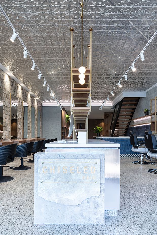 A spacious, polished salon interior viewed toward a central reception island. The room features a pale stone reception desk with a mounted “Chiseled Hair” sign, terrazzo-style flooring, dark salon chairs and styling stations along both sides, a blue-toned service counter, open shelving, plants, and a staircase leading to an upper level. A highly decorative pressed-metal ceiling, exposed track lighting, and a suspended linear pendant create a distinctive commercial fit-out with a refined industrial feel.