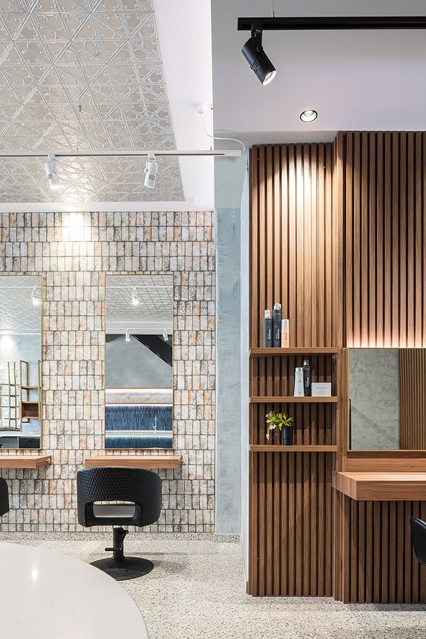 A contemporary commercial hair salon interior featuring styling stations with mirrors, black salon chairs, integrated shelving, and warm timber slatted cabinetry. The left wall is clad in pale textured brick-like tiles, while the ceiling has decorative patterned panels and track lighting. A terrazzo-style floor and neutral palette are accented by rich timber joinery and warm illumination.