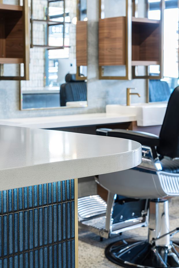 A close-up view of a stylish contemporary barbershop or salon interior. A large white rounded countertop with a textured edge projects over a dark blue vertically ribbed feature panel. Behind it are salon workstations with black leather barber chairs, mirrors, timber-toned wall cabinets, white basins, and brushed brass tapware. The space is bright and polished, with a refined commercial fit-out and strong blue, white, timber, and metallic accents.