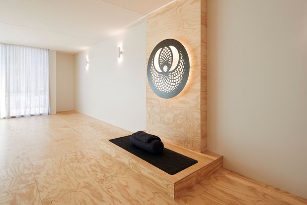 Minimal, warm-toned interior featuring a raised timber platform and integrated low bench or ledge. Pale plywood or timber surfaces continue across the floor, stepped platform, wall feature panel and built-in joinery. A large circular decorative wall artwork is backlit above a dark cushion or upholstered object, while sheer full-height curtains diffuse daylight at the far end. Recessed and discreet wall lighting creates a calm, uncluttered atmosphere.