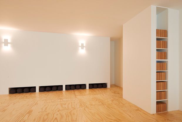 Minimalist interior room with pale walls, a warm plywood floor, recessed low-level wall niches containing dark rounded objects, simple wall sconces, and a tall open shelving unit built into the right wall. The space is sparsely furnished with clean geometric forms and warm ambient lighting.