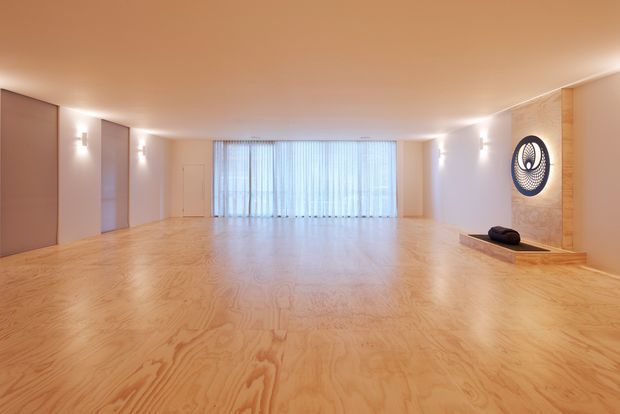 A large, minimalist open-plan interior with a warm plywood floor and matching warm-toned ceiling. Smooth pale walls are interrupted by several flush wall panels and discreet wall lighting. Full-width sheer curtains cover the rear glazing, while a timber-clad feature wall at right incorporates a shallow built-in fireplace or recessed display with dark sculptural decor. The room is empty of furniture, creating a calm, gallery-like atmosphere.