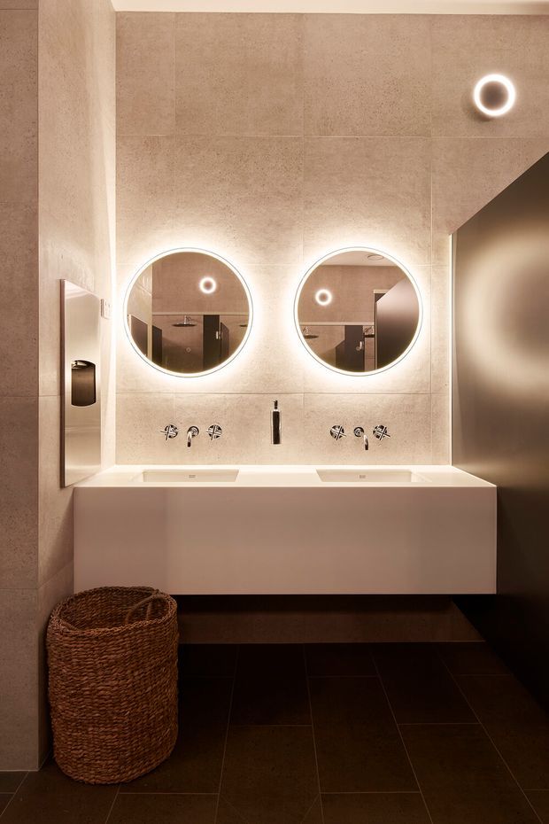 A sophisticated contemporary bathroom featuring a floating double vanity with integrated white basins, wall-mounted chrome fixtures, large beige stone-look wall tiles, dark floor tiles, and two illuminated round mirrors. Warm indirect lighting creates a refined, moody atmosphere, with a woven basket adding natural texture.