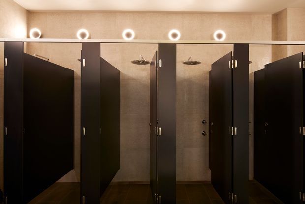 A dimly lit row of dark privacy shower cubicles in a commercial bathroom or changing-room setting. Each stall has black partition doors and walls, warm textured beige wall surfaces, dark tiled flooring, overhead round showerheads, and circular illuminated wall lights above the partitions.