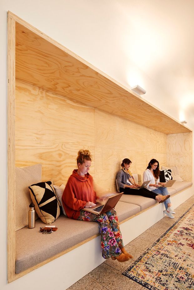 A warm, contemporary communal workspace or lounge with a long built-in upholstered bench set against a full-height plywood-lined wall and projecting plywood canopy. Three people are working on laptops, while patterned cushions and a large patterned rug add visual interest. The space has polished concrete or terrazzo-like flooring, recessed/track lighting, and a light timber palette.