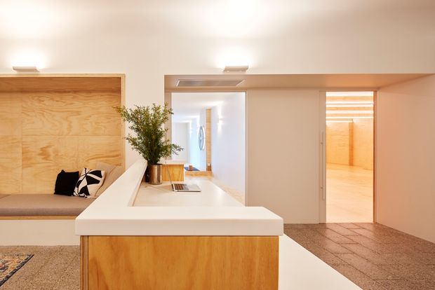 A warm, minimalist interior centered on a compact built-in timber kitchen or kitchenette island with a thick white benchtop. A timber-lined built-in seating nook with cushions is visible at left, while a white hallway and open doorway lead to another timber-accented room. The space features smooth white walls and ceiling, integrated cabinetry, recessed lighting, and a speckled hard floor.