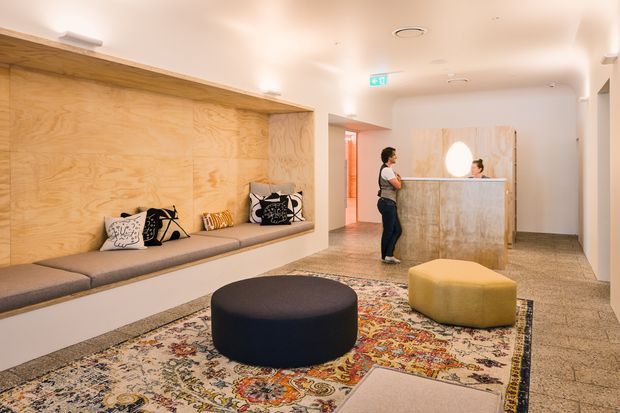 A warm, contemporary reception lobby with a built-in timber-lined bench along one wall, patterned cushions, a stone or solid-surface reception desk, terrazzo-style flooring, and a large patterned rug with rounded upholstered ottomans. A receptionist and visitor stand near the desk, while concealed and recessed lighting creates an inviting atmosphere.