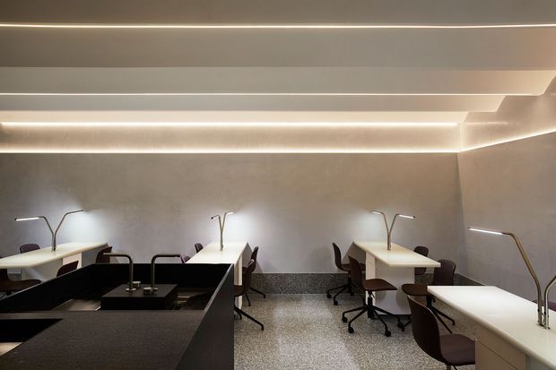 A refined contemporary commercial workspace with multiple shared desks arranged along the walls, ergonomic office chairs, slim task lamps, integrated linear cove lighting, and a dark central kitchenette or service counter. The room has softly textured grey walls, a speckled terrazzo-like floor, and a calm, softly illuminated atmosphere.
