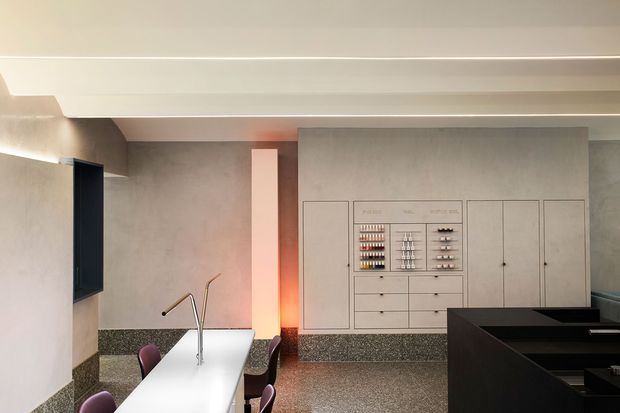 Contemporary open-plan kitchen with a long white island, integrated sink and tap, dark cabinetry, terrazzo flooring, and a full-height built-in storage wall. The space has a refined neutral palette with warm concealed lighting and a partially visible adjacent dining or living area.