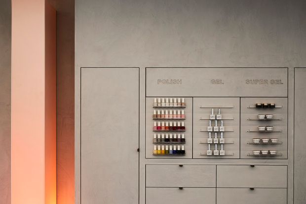 A close-up of a refined built-in display and storage wall with a warm textured plaster finish. The central open display section presents rows of small product samples or containers, flanked by a tall concealed cabinet and multiple wide drawers. Slim dark metal pulls and fine black outlines create a precise, minimalist composition, with warm ambient lighting from the left.