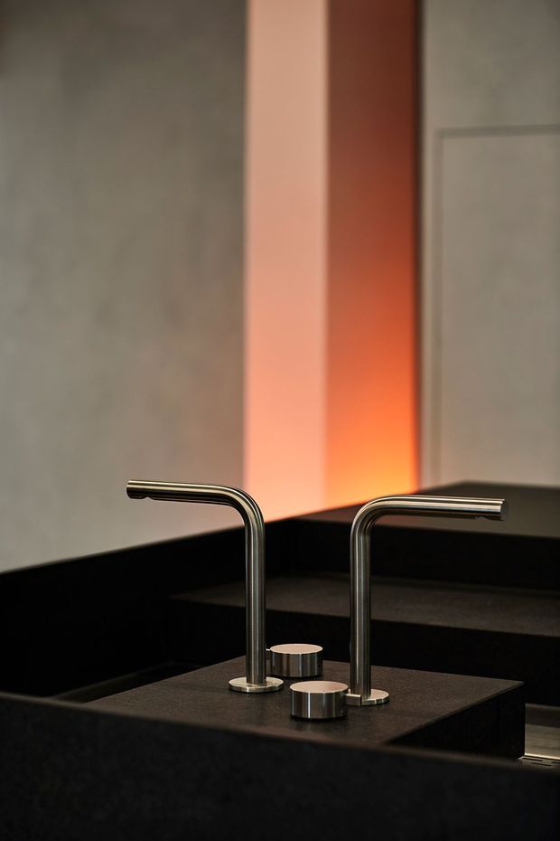 Close-up architectural detail of a dark, minimalist benchtop with two tall brushed-metal gooseneck faucets and cylindrical controls. The surrounding cabinetry and surfaces are predominantly black, with a warm orange illuminated vertical recess creating contrast against pale wall panels.
