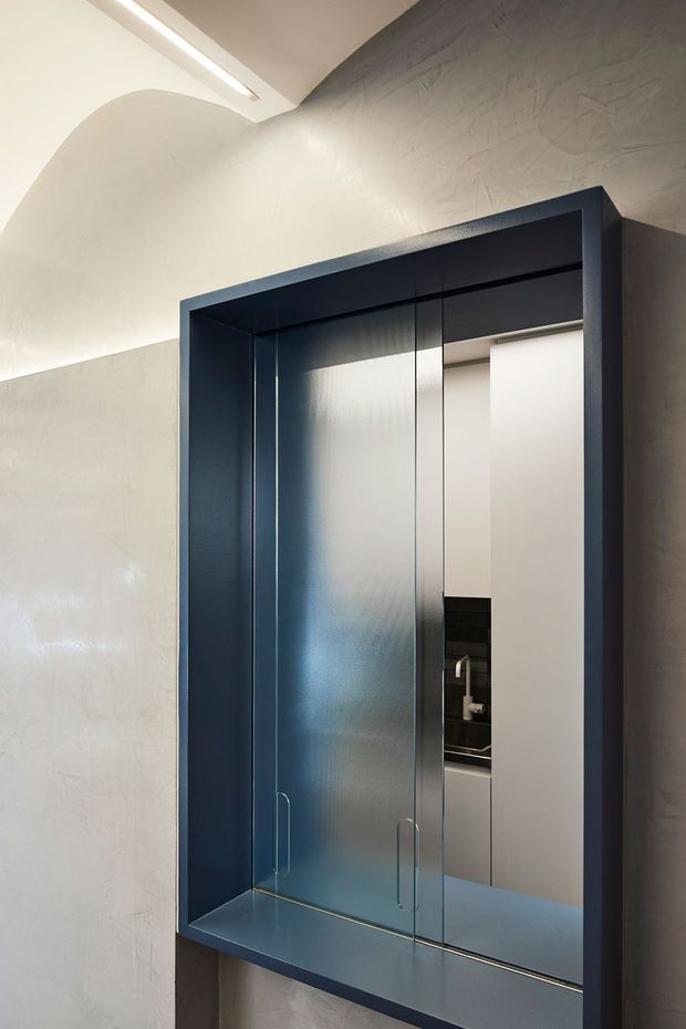 A close-up architectural interior detail showing a dark blue-black framed glass door or shower enclosure set into textured pale plaster walls. The reflective glazing reveals a compact bathroom vanity and basin beyond. A linear recessed ceiling light and softly curved plaster transition add sculptural detail to the minimalist space.