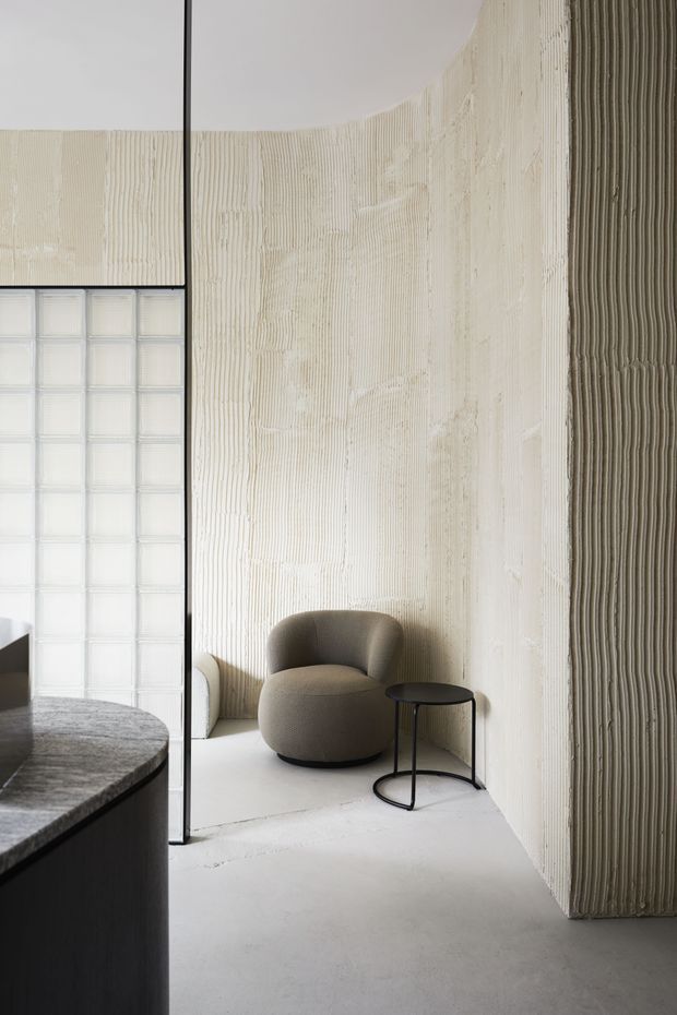 A minimalist interior corner with a rounded textured plaster wall, pale concrete-like floor, and a sculptural taupe lounge chair beside a small black side table. A black-framed glass-block partition and dark curved built-in counter edge appear on the left. The space has soft natural light, monochromatic neutral tones, and strong vertical ribbed wall texture.