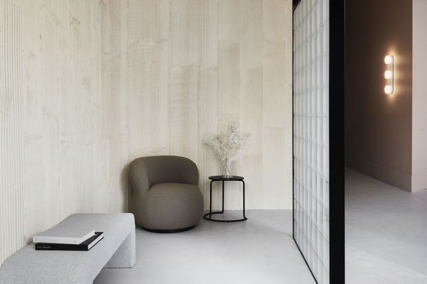 Minimalist interior lounge or seating area with a sculptural upholstered armchair, curved light-grey concrete or stone-like coffee table, small black metal side table, and sparse dried-branch styling. The room features a textured off-white wall, pale grey floor, and a translucent glass-block partition opening toward a darker adjacent space with a vertical wall light.