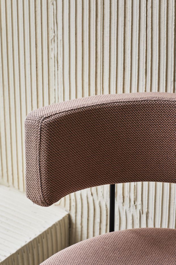 Close-up product-style image of a curved upholstered chair with a rounded brown textured fabric backrest and seat, set against a vertically ridged light timber or plaster feature wall. A pale sculptural side table is partially visible.