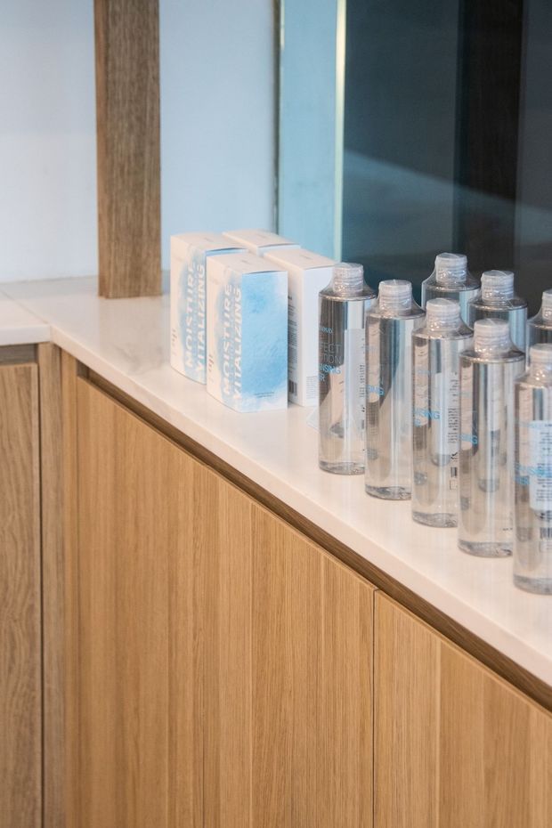 Close-up of a refined built-in timber vanity or display cabinet with a pale stone countertop, showcasing neatly arranged clear cosmetic or skincare bottles and boxed products. A large mirror or dark reflective panel is visible behind the counter, with light timber joinery and minimal detailing creating a clean contemporary appearance.
