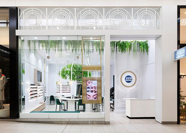 Bright contemporary retail beauty salon frontage viewed from a shopping-centre corridor. The predominantly white storefront has extensive clear glazing, a decorative geometric metal grille above, hanging greenery, a white reception desk, display shelving, mirrors, and salon workstations visible inside.