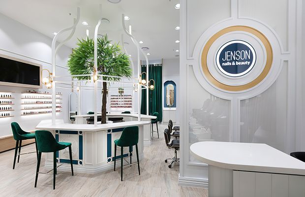 Bright, refined nail and beauty salon interior with white decorative wall panelling, pale timber-look flooring, curved white manicure counters, teal upholstered stools, extensive product shelving, and a central indoor tree feature beneath sculptural pendant lighting. A circular Jensen nails & beauty sign forms a prominent wall feature.