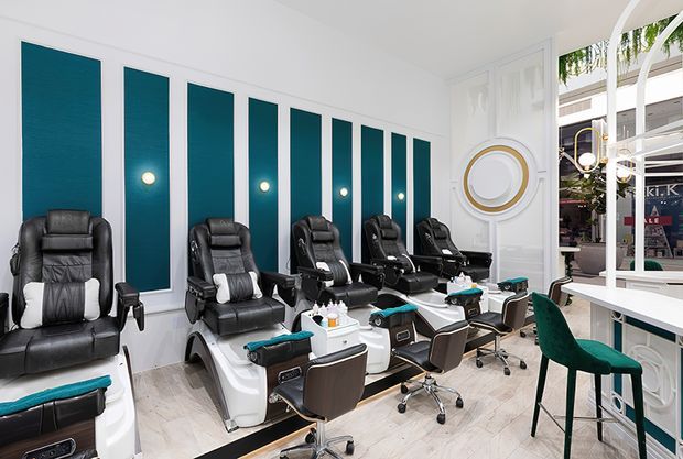Bright, high-end nail and pedicure salon interior with multiple black reclining pedicure chairs and white foot spa basins arranged along a wall. The room features repeated tall teal wall panels with integrated round lights, white decorative mouldings, a circular wall feature, pale marble-look flooring, and a white-framed glazed frontage with green velvet bar stools. The overall design is polished, contemporary, and luxe with strong teal-and-white contrast.