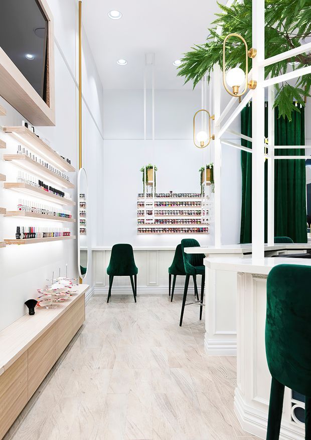 Bright, narrow nail salon interior with white walls and built-in display shelving filled with nail polish bottles. The space features pale timber-look flooring, white decorative wall mouldings and partitions, deep green velvet chairs, mirrored surfaces, indoor plants, and elegant brass-and-glass wall sconces. The overall design is refined, bright, and high-contrast with a boutique commercial fit-out.