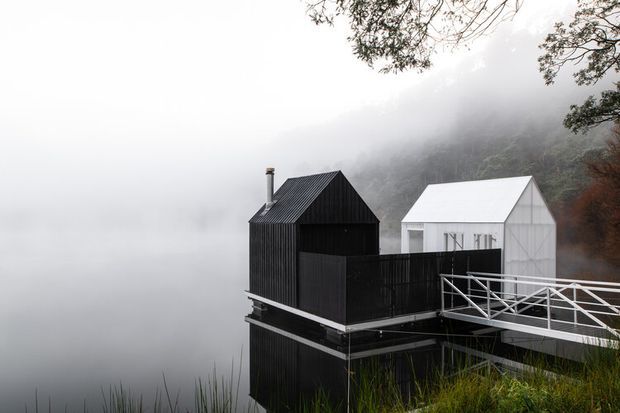 A striking pair of small contemporary structures elevated above a misty waterfront or wetland setting. The foreground building is clad in dark vertical timber with a steep gable roof, chimney, enclosed upper deck, and raised supports. A smaller white gabled building stands behind it, connected by a white metal pedestrian bridge. Dense fog, reeds, and wooded slopes create a secluded, atmospheric setting.