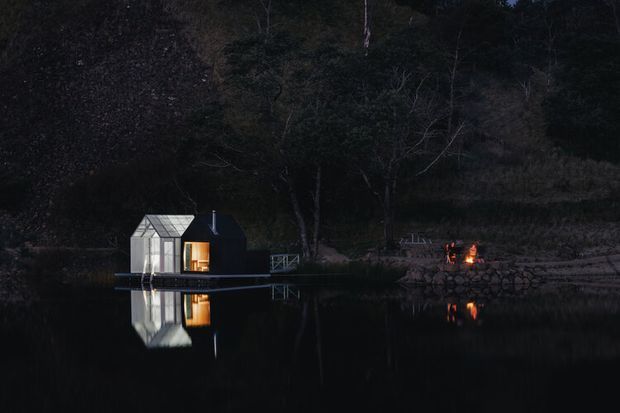A small modern cabin or pavilion sits beside a still, dark lake at night, surrounded by dense forest. The compact structure has a sharply pitched, gabled roof, dark exterior walls, and warm light glowing through its windows and doorway. The building and surrounding trees are reflected in the water, while a small fire or illuminated outdoor feature is visible on the shore to the right.