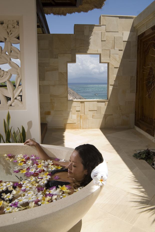 A sunlit luxury bathing courtyard featuring a freestanding stone bathtub filled with flower petals, surrounded by pale natural-stone walls and flooring. A decorative carved screen and timber door frame the space, while a geometric opening reveals the ocean beyond. The setting has an open-air tropical resort character.