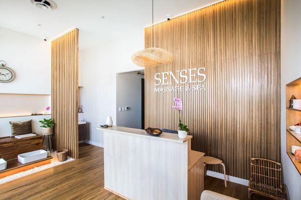 Bright, warmly styled spa reception area with a light timber reception desk, vertical timber feature wall and screening, white walls, wood-look flooring, woven pendant lighting, built-in shelving, and a small seating area. The space is identified by visible signage as a massage and spa business.
