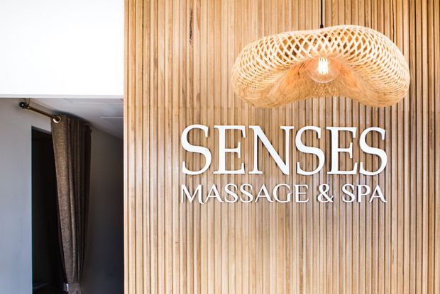 Interior branding wall for a massage and spa, featuring a vertical timber slat feature wall, large white raised lettering reading “Senses Massage & Spa,” and a woven sculptural pendant light. A curtained doorway is visible at the left edge.