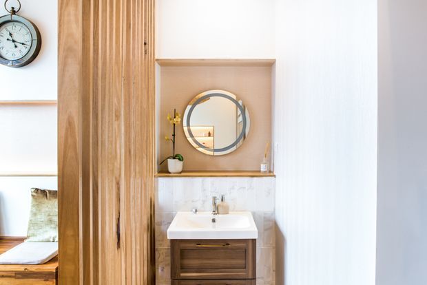 Bright, compact bathroom vanity area featuring a timber floating cabinet, white rectangular basin, brass tapware, pale tiled splashback, and a round framed mirror set within a recessed wall niche. Vertical timber screening partially separates the bathroom from an adjacent sleeping or seating area visible at left. The space has warm natural materials and clean contemporary detailing.