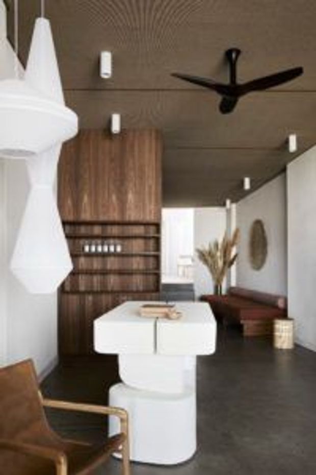 A contemporary open-plan kitchen and dining area with a sculptural white island, dark timber cabinetry and integrated shelving, a white pendant light, dark flooring, and a wood-panelled ceiling with a black ceiling fan. A lounge area with a brown sofa and decorative styling is visible beyond the kitchen.