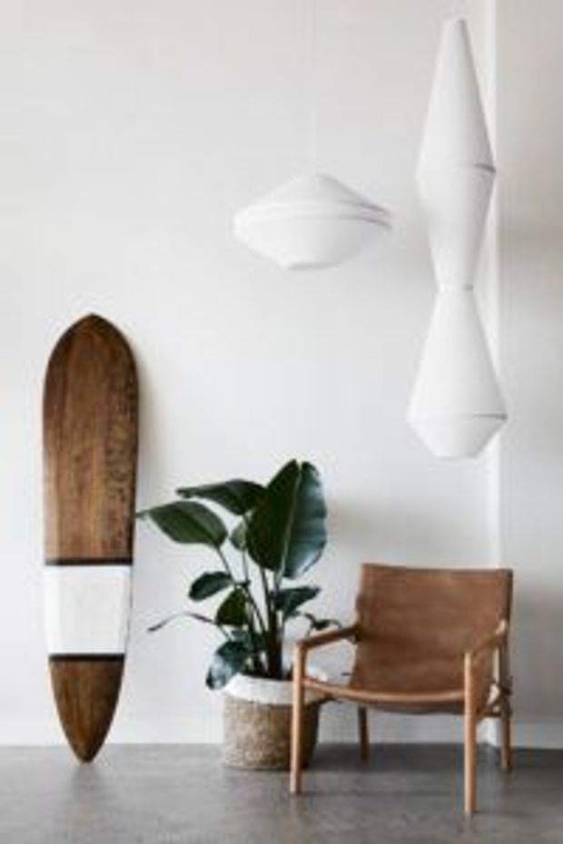 Minimalist interior vignette with a white wall, grey concrete-like floor, a suspended white sculptural pendant light, a brown leather-and-timber armchair, a potted green plant, and a vertically mounted wooden surfboard-shaped decorative object.