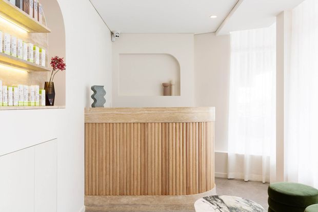 Bright, softly styled reception or living space featuring a curved fluted timber-clad counter, integrated illuminated shelving with books and decor, an arched wall niche, sheer floor-to-ceiling curtains, and neutral finishes. The room has a clean contemporary design with warm natural timber accents.