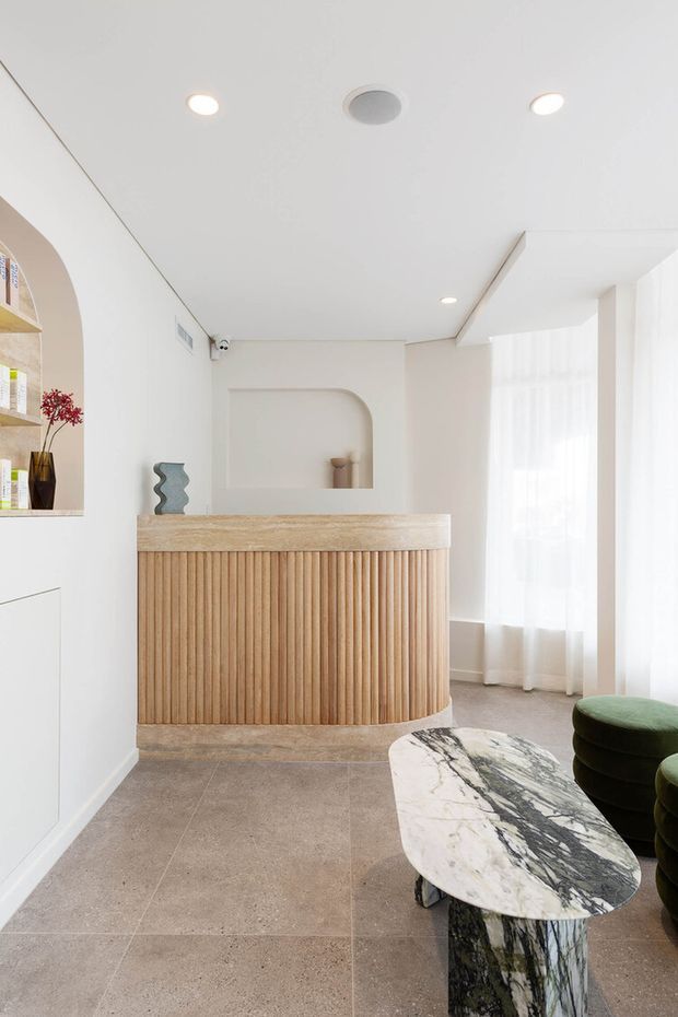 Bright, refined reception or entry area with a fluted timber-fronted curved counter, pale stone benchtop, arched wall niche, integrated shelving, sheer floor-to-ceiling curtains, and a sculptural marble coffee table with green upholstered seating. The space has a clean contemporary fit-out with warm natural materials and soft neutral tones.
