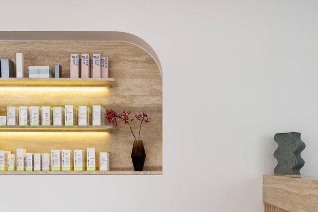 Minimalist interior detail featuring a built-in, rounded-edge display shelving unit clad in warm beige stone or stone-look material, with integrated warm LED shelf lighting and neatly arranged product boxes. A matching stone console or pedestal with a sculptural dark decorative object appears at right against a plain light wall.
