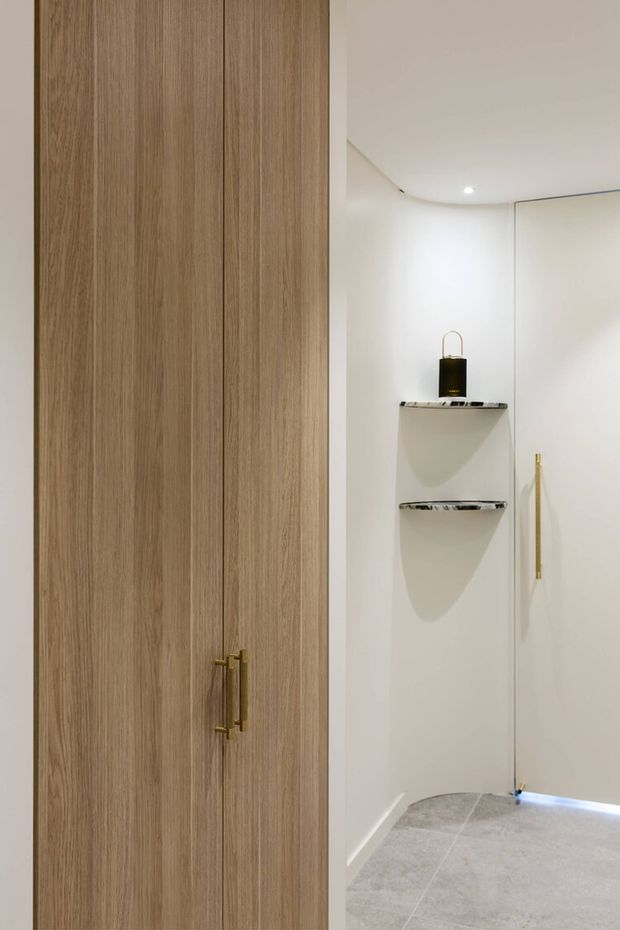 A minimalist built-in storage or wardrobe area with tall vertical light-wood cabinet doors and brass handles. Adjacent white recessed shelving features dark stone or marble-edged shelves, while a full-height mirrored door with a slim brass pull reflects the clean white interior. The space has pale grey stone-look tiled flooring, curved ceiling detailing, and discreet recessed lighting.